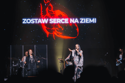 Na scenie koncertowej dwie osoby śpiewają, jedna trzyma mikrofon. W tle duży ekran z grafiką czerwonego serca i napisem „ZOSTAW SERCE NA ZIEMI”. Po lewej perkusista za przezroczystą osłoną, po prawej gitarzysta. Scena ciemna, oświetlona reflektorami.
