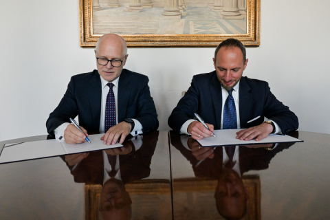 Dwóch mężczyzn w garniturach siedzi przy stole i podpisuje dokumenty.