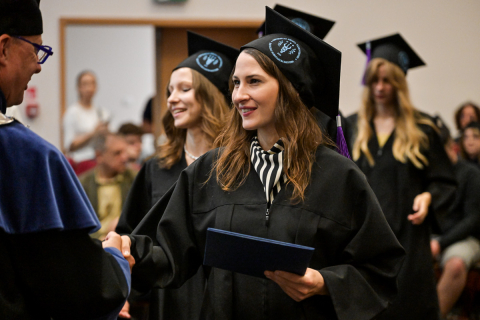 Absolwentka w czarnej todze i birecie z fioletowym chwostem odbiera dyplom. Prawą dłonią ściska dłoń osoby wręczającej. W tle widoczni są inni absolwenci i goście. 