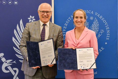 Mężczyzna w szarym garniturze i kobieta w różowym garniturze prezentują dokumenty w otwartych teczkach. W tle dwa banery z logotypami Warszawskiego Uniwersytetu Medycznego.