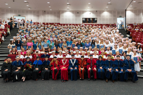 Uroczystość czepkowania absolwentów pielęgniarstwa i położnictwa w audytorium. W pierwszym rzędzie siedzą osoby w togach akademickich - kadra naukowa. Za nimi stoją absolwenci w różnokolorowych strojach, na głowach mają czepki pielęgniarek i położnych, a w rękach dyplomy.