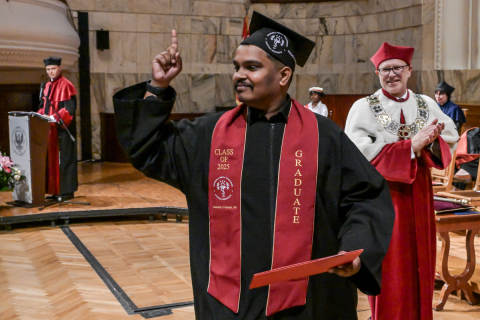 Absolwent w czarnej todze i birecie z czerwonym szalikiem z napisem „CLASS OF 2023” i „GRADUATE” trzyma dyplom w lewej ręce. Prawą rękę unosi z wyprostowanym palcem wskazującym. W tle osoby w togach, jedna przemawia przy mównicy.