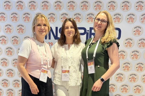 Trzy kobiety stojące przed ścianką z logo ‘INSPIRED’ oraz wzorem przedstawiającym dwie dłonie trzymające serce. Każda z kobiet ma zawieszony na szyi identyfikator.