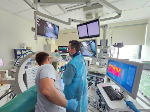 W szpitalnym pomieszczeniu medycznym odbywa się procedura diagnostyczna lub zabiegowa. Na pierwszym planie widocznych jest dwóch pracowników – jeden ubrany w niebieski fartuch ochronny i rękawiczki, drugi w codziennym stroju. W tle znajdują się liczne monitory wyświetlające obrazy medyczne, takie jak zdjęcia rentgenowskie. 