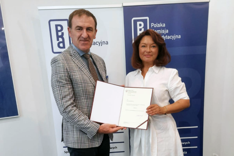 Dwie osoby stoją przed dwoma roll-upami z napisem „Polska Komisja Akredytacyjna”. Wspólnie trzymają rozłożoną teczkę z dokumentem. Mężczyzna po lewej ma szarą marynarkę w kratę. Kobieta po prawej, prof. Aneta Nitsch-Osuch, nosi białą koszulę. Obie osoby uśmiechają się do zdjęcia.