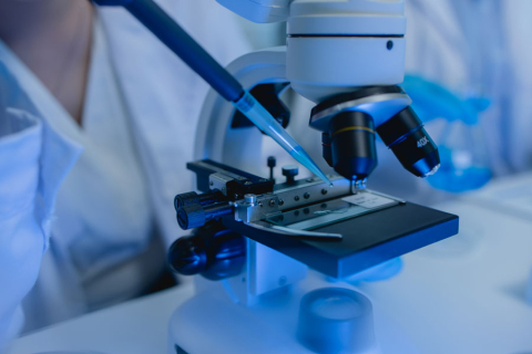 Mikroskop laboratoryjny z próbką na szkiełku. Nad próbką unosi się pipeta trzymana przez osobę poza kadrem. W tle widoczne fragmenty białych fartuchów. Całość oświetlona niebieskim światłem.