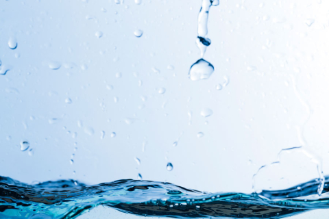 Krople wody spadają na falującą powierzchnię wody. Tło jest jasnoniebieskie. Na pierwszym planie widać rozpryski i fale. Krople mają różne rozmiary i tworzą dynamiczny efekt.