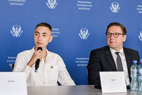 Młody człowiek w białej koszuli z haftowanym logo na piersi trzyma mikrofon. Obok niego siedzi mężczyzna w ciemnym garniturze, białej koszuli i krawacie. Obaj siedzą przy stole podczas konferencji prasowej. 