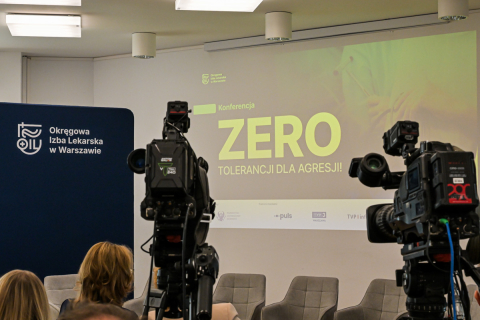 Ekran z napisem zero tolerancji dla agresji i dwie kamery. Obok ekranu widać niebieską ściankę z białym logotypem Okręgowej Izby Lekarskiej. 