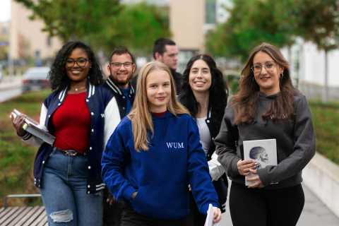 Studenci WUM: cztery kobiety i dwóch mężczyzn. Studenci idą chodnikiem. Uśmiechają się. Są ubrani w bluzy i jeansy. Dziewczyna na pierwszym planie ma granatową bluzę i jasne włosy. Dziewczyna po lewej ciemne włosy, okulary i czerwoną koszulkę. Dziewczyna po prawej ma szarą bluzę, okulary i biały notes z grafiką modelu czaszki. Pozostałe osoby są mniej widoczne. W tle widać budynki, zielone drzewa i krzewy.