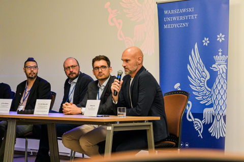 Czterech ubranych elegancko mężczyzn. Siedzą przy stole. Mężczyzna po prawej trzyma w ręce mikrofon, przemawia. Pozostali spoglądają w jego stronę. Za nimi niebieski baner z logo WUM - białym orłem.