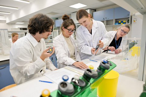 Grupa osób w białych fartuchach laboratoryjnych w trakcie zajęć. Część osób ma w rękach pipety do miareczkowania. Przed nimi na blacie leżą materiały edukacyjne.