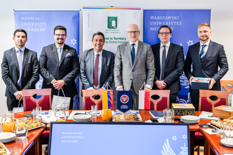 Sześciu stojących mężczyzn. Wszyscy są elegancko ubrani. Uśmiechają się do zdjęcia. W tle wiszą trzy banery. Po lewej i po prawej niebieskie banery z logo WUM, w środku biały baner uniwersytetu z Kolumbii.