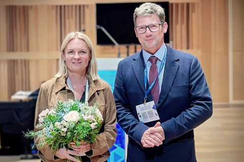 Dwie elegancko ubrane osoby. Kobieta po lewej, prof. Urszula Ambroziak, trzyma bukiet kwiatów. Mężczyzna po prawej to prof. Przemysław Witek. Oboje uśmiechają się do zdjęcia. Stoją w pomieszczeniu wyłożonym drewnem, za nimi widać ekran i mównicę.