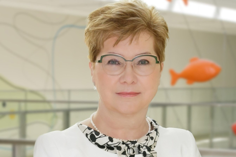 Zbliżenie twarzy kobiety w okularach, o krótkich, jasnych włosach, ubrana w biały strój,. W tle nowoczesne wnętrze z abstrakcyjną dekoracją ścienną i pomarańczową ozdobą w kształcie ryby zawieszoną pod sufitem