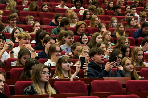 Grupa młodych kobiet i mężczyzn siedzi w audytorium, zwrócona w stronę sceny. Wiele osób trzyma telefony komórkowe, robiąc zdjęcia lub nagrywając filmy