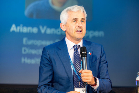 An elegantly dressed middle-aged man, Dr. Wojciech Feleszko, speaks at a forum. He holds a microphone in his hand. Behind him is a multimedia screen.
