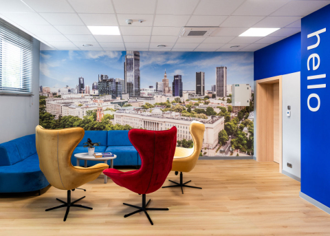 Jasny pokój biurowy z drewnianym parkietem i dużą fototapetą miasta z wieżowcami. Cztery fotele — dwa żółte, jeden czerwony i jeden niebieski — stoją wokół okrągłego stolika. Po lewej stronie znajduje się niebieska kanapa. Na prawej ścianie widnieje napis „hello” na niebieskim tle. Przez duże okno z żaluzjami wpada światło dzienne.