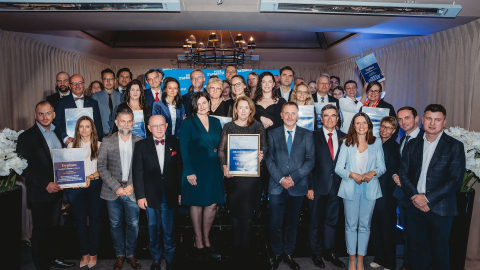 Grupa elegancko ubranych osób pozuje do wspólnego zdjęcia. Część z nich trzyma w rękach oprawione dyplomy.