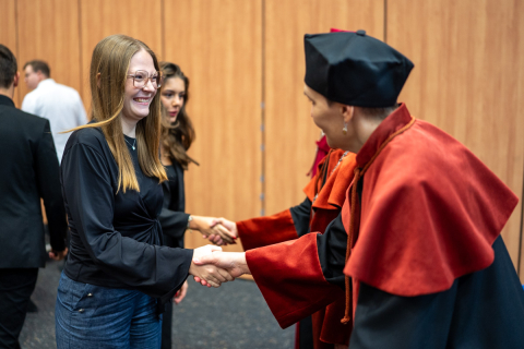 Prodziekan ds. nauczania na kierunku farmacja, w czarno-pomarańczowe todze, podaje rękę młodej dziewczynie - studentce, w czarnej bluzce i jeansach. Studentka szeroko się uśmiecha. Obok stoją dwie osoby w togach. Na dalszym planie widać też inne osoby.