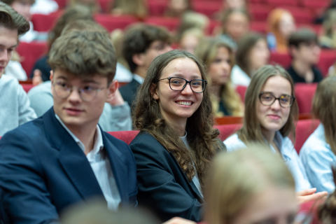Grupa młodych osób siedząca na czerwonych fotelach. Wśród nich dwie młode dziewczyny patrzą prosto w obiektyw i uśmiechają się do zdjęcia.