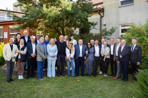 CopilotHere is the English translation of your text: A group of women and men is standing on the grass in front of a building, posing for a group photo. In the background, there is a tree and parts of the building with windows. Some people are dressed in elegant suits, others in casual clothes.