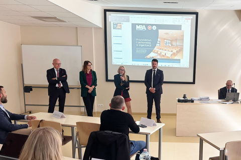 A group of people in a lecture hall. Four people are standing in the middle, one of them—a woman—has a microphone and is speaking. Behind them, a multimedia screen can be seen.