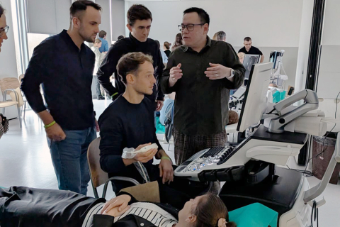 Six people in a bright room with large windows. One person is lying on a treatment bed covered with green fabric, and another is sitting next to them holding an ultrasound probe. Three people are standing nearby watching the ultrasound screen, and one person in the background is working at another station. Medical accessories are lying on the desk next to the machine. 