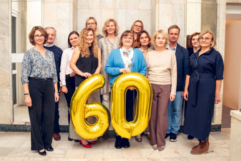 Grupa kobiet i mężczyzn stoi w jasnym pomieszczeniu z marmurowymi ścianami. W centrum dwie kobiety trzymają duże, złote balony w kształcie liczby „60”.