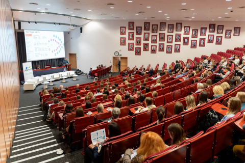 Sala wykładowa z czerwonymi krzesłami, sceną z białymi fotelami i ekranem projekcyjnym. Na prawej ścianie portrety w czerwonych ramkach. Uczestnicy skupieni na prezentacji.