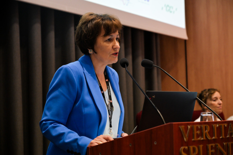 Kobieta w niebieskiej marynarce i białej bluzce przemawia na konferencji, stojąc za drewnianą mównicą