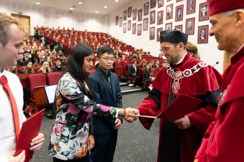 Dziekan, ubrany w czerwoną togę i czarny biret oraz prorektor, ubrany w czerwoną togę, składają gratulację nowo przyjętym studentom. W tle widać pełną aulę.