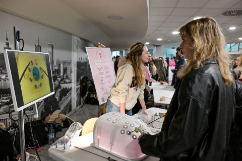 Dwie osoby stoją przy stole z symulatorem medycznym w kolorze białym i różowym. Jedna osoba w jasnej bluzie obsługuje stanowisko, druga w czarnej kurtce patrzy na ekran z kolorowym obrazem narządów. W tle widać plakat z instrukcjami i grupę ludzi przy innych stoiskach.