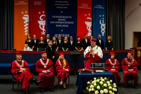 Sześć osób w czerwonych togach i biretach z insygniami rektorskimi na scenie auli. Jedna z nich, rektor, stoi i przemawia do mikrofonu. Pozostali siedzą. W tle widać chór w czarnych strojach oraz kolorowe banery z nazwami wydziałów. Na stole przed mównicą leży bukiet białych kwiatów. Scena przedstawia inaugurację roku akademickiego 2025/2026