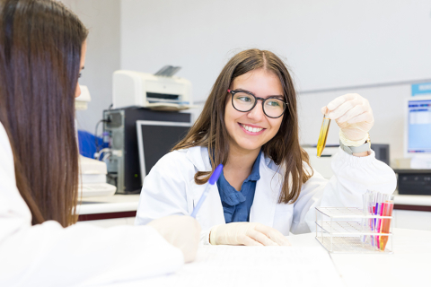 Two young women wearing white coats are sitting at a table in a laboratory. One of them, facing the camera, is holding a vial with a preparation, while the other, with her back to the camera, is writing something in a notebook. Laboratory equipment can be seen behind them.