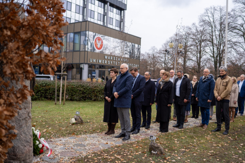 Grupa osób stoi przed kamiennym obeliskiem. Pod obeliskiem leży wieniec z kwiatów. W tle widać zieleń oraz szaro-brązowy budynek.