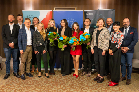 A group of people are standing in a conference room in front of three roll-ups with logos. Three people in the middle are holding large bouquets of blue and orange flowers.