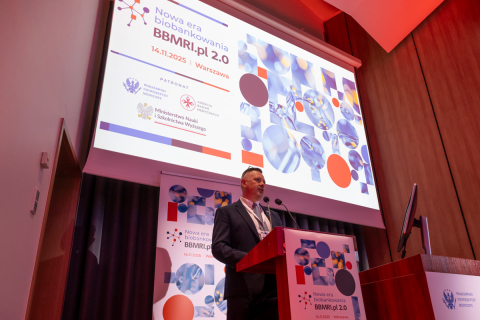 A man at the podium during a conference. Behind him is a multimedia screen displaying the conference topic and additional information.
