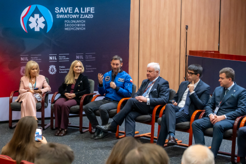 Sześć osób siedzi w półokręgu na krzesłach podczas panelu dyskusyjnego. W tle znajduje się granatowa ścianka z napisem ‘SAVE A LIFE – Światowy Zjazd Polonijnych Środowisk Medycznych’. Jedna osoba trzyma w ręce mikrofon i mówi - to Sławosz Uznański-Wiśniewski. W tle widać granatową ściankę z logo Save a Life.