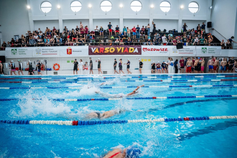 Zawody pływackie. Na pierwszym planie widać basen oraz płynących w nim zawodników. W oddali widać trybuny, na których siedzą ludzie.
