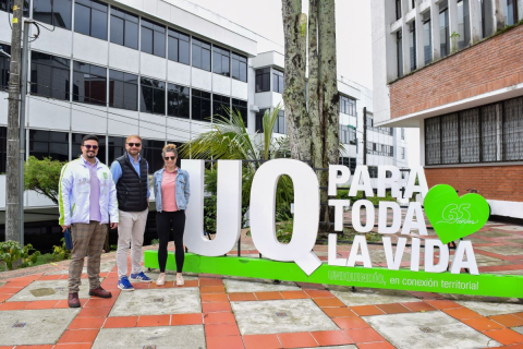 Three people standing next to a large white installation with the words “UQ PARA TODA LA VIDA” and a green heart with the number “65 años.” 
