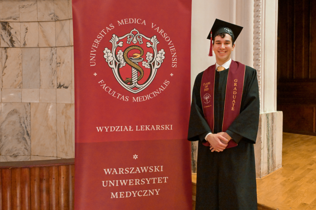 Absolwent Warszawskiego Uniwersytetu Medycznego w todze i birecie stoi obok czerwonego baneru z herbem uczelni i napisami: „UNIVERSITAS MEDICA VARSOVIENSIS”, „FACULTAS MEDICINALIS”, „WYDZIAŁ LEKARSKI”, „WARSZAWSKI UNIWERSYTET MEDYCZNY”. Ma bordową szarfę z napisem „GRADUATE” i rocznikiem. Tło to marmurowa ściana i drewniana podłoga.